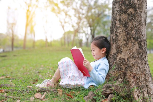 Little girl reading a book at the park with her back against a tree