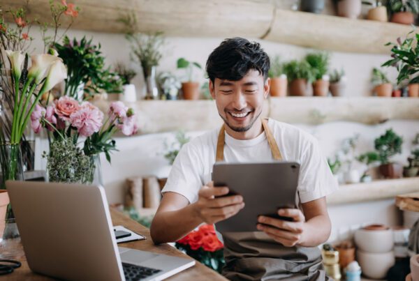 Male florist, owner of small business flower shop, using digital tablet while working on laptop to check inventory, take orders, selling products online. Daily routine of running a small business with technology.