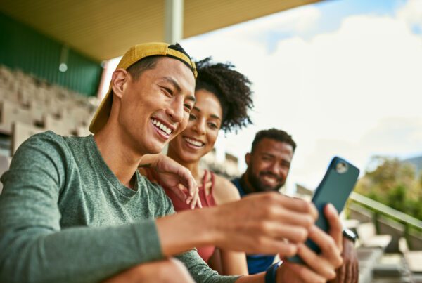 Three people outdoors at a sporting event smiling at a phone.