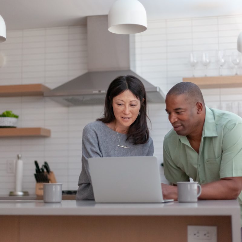 Husband and wife in their kitchen using a laptop computer to pay bills online and manage their budget.