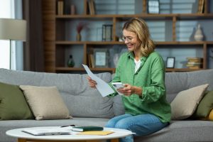 An elderly woman received good news, financial deal, loan repayment, house sale, pension, subsidy. She is sitting on the sofa at home, holding the phone and documents in her hands. Checks accounts.