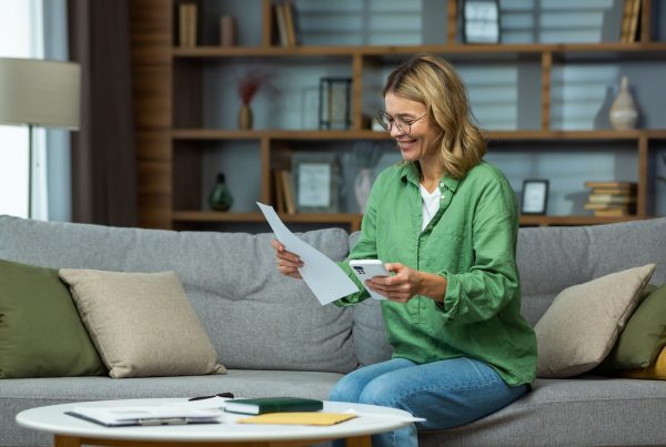 An elderly woman received good news, financial deal, loan repayment, house sale, pension, subsidy. She is sitting on the sofa at home, holding the phone and documents in her hands. Checks accounts.