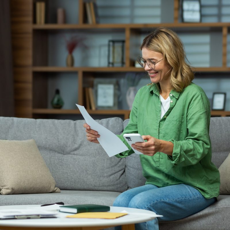 An elderly woman received good news, financial deal, loan repayment, house sale, pension, subsidy. She is sitting on the sofa at home, holding the phone and documents in her hands. Checks accounts.