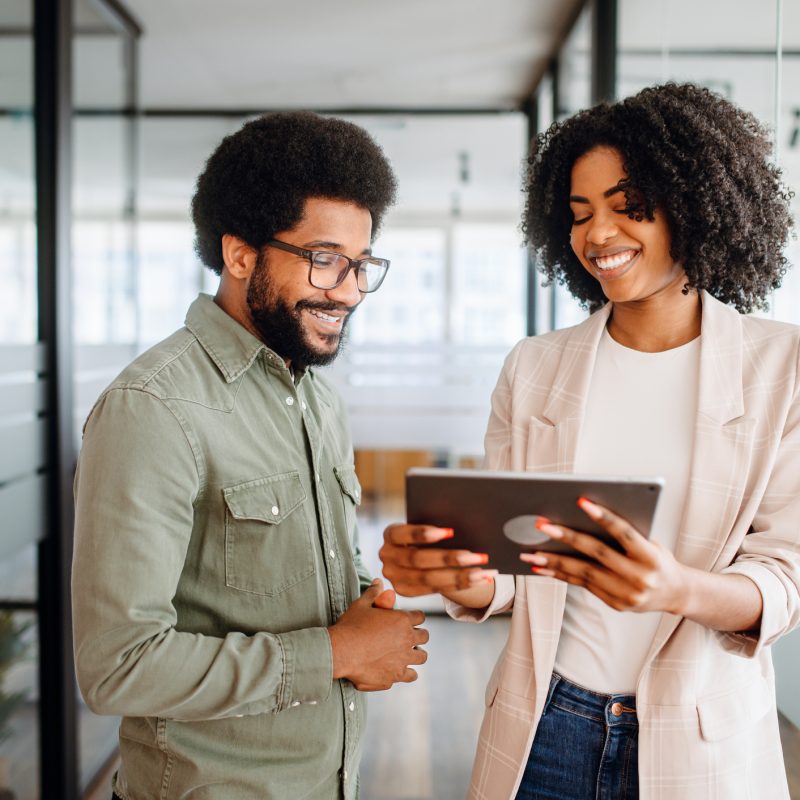 Two cheerful colleagues are sharing a tablet screen, the woman pointing at the screen, a man watching with interest, collaborative and friendly work atmosphere. Team interaction and knowledge exchange
