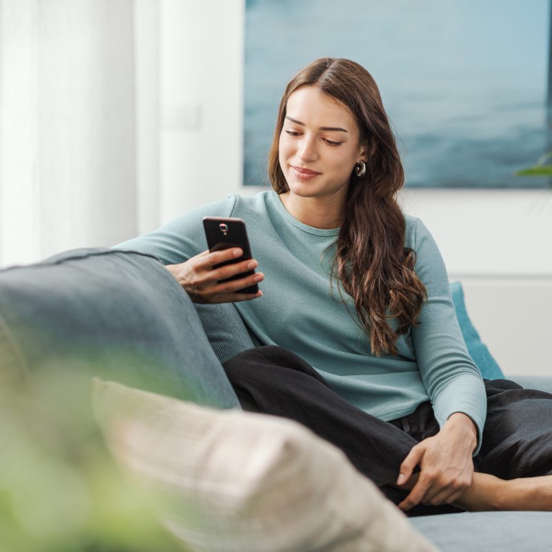 Happy young woman sitting on the couch at home and chatting with her smartphone, she is social networking and using mobile apps